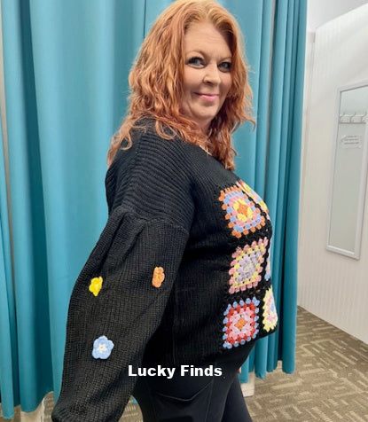 Person wearing a black cardigan with colorful patchwork patterns in front of blue curtains.